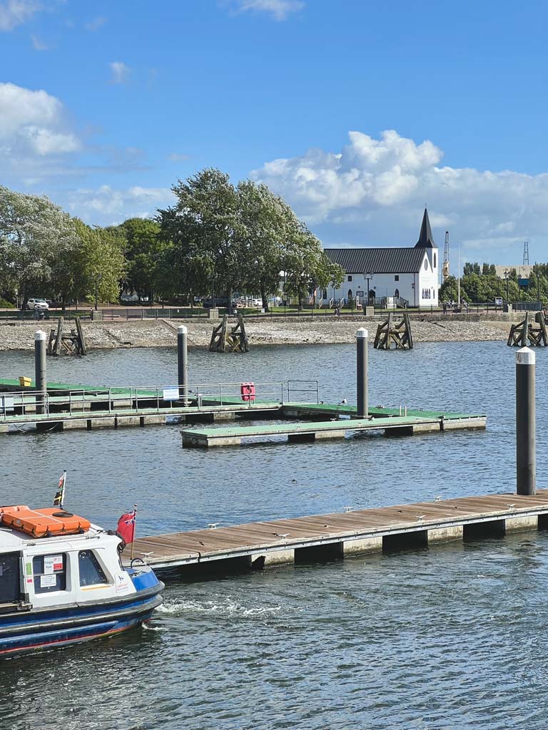 A jetty over blue water with a white church in the distance and a boat in the foreground